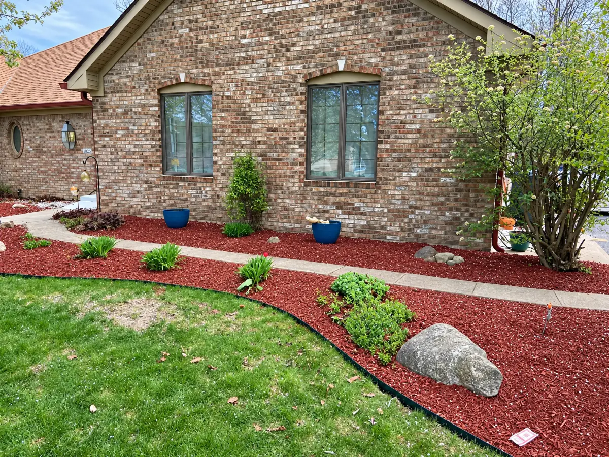 Brick home exterior with red-mulched garden beds, small plants, shrubs, decorative rocks, and blue pots along a concrete walkway and lawn.