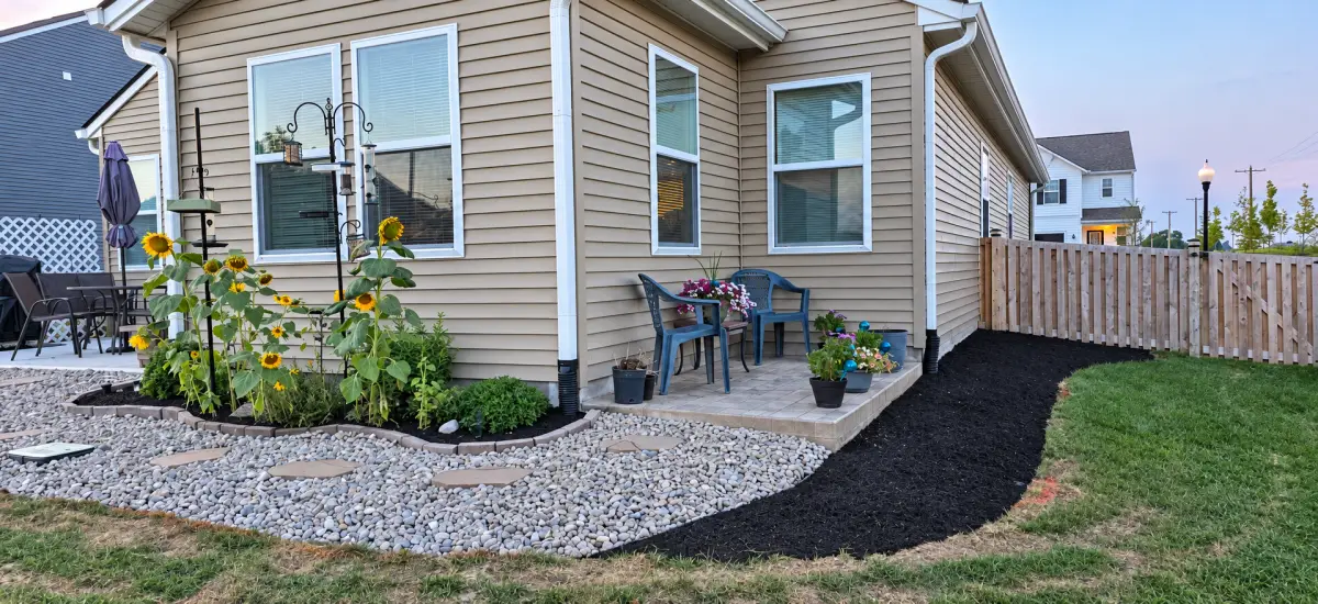 Backyard landscaping beside a house, with a curved dark mulch bed, decorative river rock, stepping stones, sunflowers, and a small patio with chairs and potted flowers.
