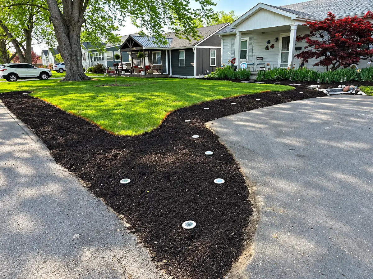 Residential front yard with a green lawn, curved dark mulch beds, round ground lights, mature trees, and planting beds beside a porch and driveway.
